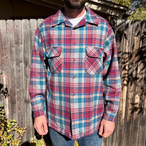May include: A long-sleeved plaid shirt in a classic style. The shirt features a pattern of red, teal, and beige squares. It has two chest pockets and a button-down front. The shirt is made of a soft, possibly flannel, material.