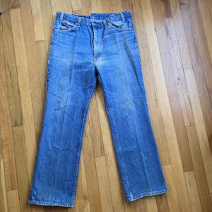 May include: Light blue denim jeans with a classic five-pocket design. The jeans have a straight leg cut and a button and zipper closure. The denim has a faded appearance, suggesting wear and washing. The jeans are laid flat on a wooden surface.