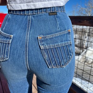 May include: A pair of blue denim jeans with a high waist and a unique pocket design. The jeans have a stitched waistband and the Wrangler brand name is visible on the back.