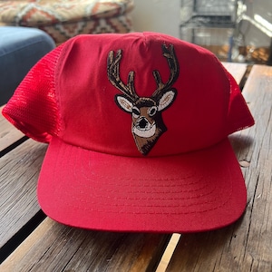 May include: A red trucker hat with a mesh back and a curved brim. The hat features an embroidered deer head with brown and tan details. The hat is sitting on a wooden surface.