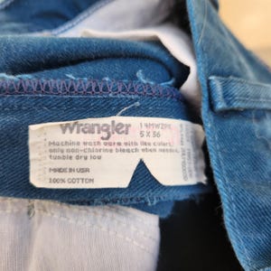 May include: Close-up of blue denim jeans with a white tag. The tag reads "Wrangler" and includes washing instructions and size 5 x 36. The jeans are made of 100% cotton and made in the USA.