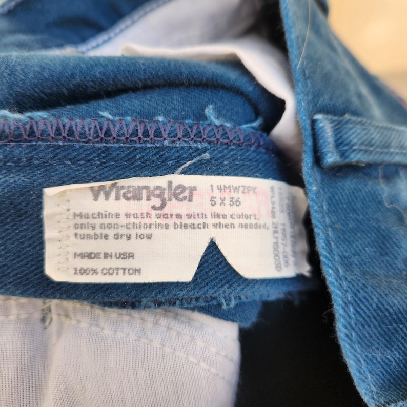 May include: Close-up of blue denim jeans with a white tag. The tag reads "Wrangler" and includes washing instructions and size 5 x 36. The jeans are made of 100% cotton and made in the USA.