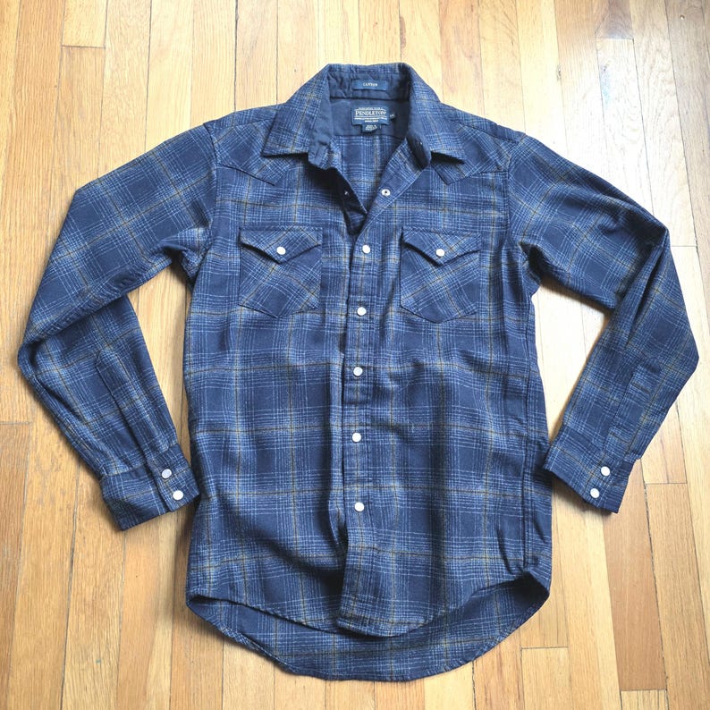 May include: A blue plaid button-up shirt with long sleeves. The shirt has two chest pockets and white buttons. The fabric has a pattern of blue and tan squares. The shirt is made of a soft, comfortable material.