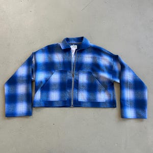 Könnte beinhalten: Blau karierte Jacke mit durchgehendem Reißverschluss und zwei Vordertaschen. Die Jacke hat einen klassischen Kragen und lange Ärmel. Das Karomuster umfasst Blau-, Weiß- und Grautöne. Die Jacke ist etwa 61 cm lang.