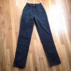 May include: Dark wash denim jeans with a high waist and straight leg. The jeans have a button closure and a small logo detail near the front pocket. The pants are laid flat on a wooden surface.