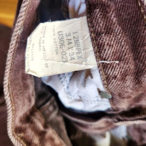 Pode incluir: Close-up de jeans de ganga castanhos com uma etiqueta branca. A etiqueta diz "12NBPEX 3/4 K 34 U306-021 100% COTTON MADE IN MEXICO OF U.S. FABRIC". Os jeans s&atilde;o feitos de algod&atilde;o.