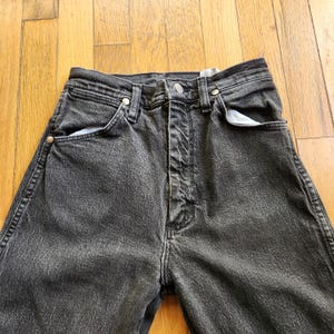 May include: Close-up of black denim jeans. The jeans have a button and zipper closure, with front and back pockets. The denim is faded, giving the jeans a worn look. The jeans are on a wooden surface.