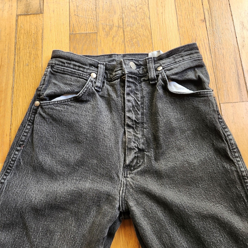 May include: Close-up of black denim jeans. The jeans have a button and zipper closure, with front and back pockets. The denim is faded, giving the jeans a worn look. The jeans are on a wooden surface.