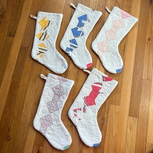 Könnte beinhalten: Fünf gesteppte Weihnachtsstrümpfe mit verschiedenen geometrischen Mustern in Gelb, Blau, Rosa und Rot. Jeder Strumpf ist weiß mit einem gesteppten Design und einer Schlaufe zum Aufhängen. Die Strümpfe sind auf einer Holzoberfläche angeordnet.
