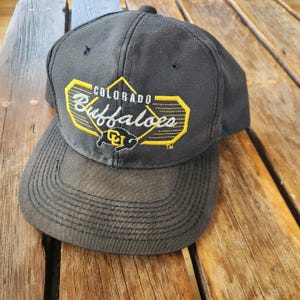 Puede incluir: Gorra de béisbol negra con el texto "Colorado Buffaloes" en amarillo y blanco, y un logotipo de búfalo. La gorra tiene una visera curva y una corona estructurada. Fabricada con tejido resistente.