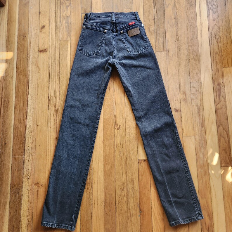 May include: Dark wash denim jeans with a classic five-pocket design. The jeans feature a red logo patch and a brown leather brand patch on the back. The jeans are laid flat on a wooden surface.