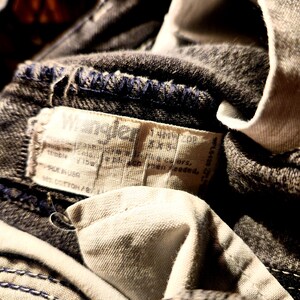 May include: Close-up of a pair of dark denim jeans with a visible Wrangler label. The label is rectangular and made of a light-colored fabric, with text in black. The jeans have blue stitching and a white fabric lining.