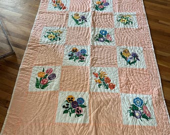 Op de afbeelding: Een vintage quilt met een roze achtergrond en witte vierkanten met geborduurde bloemenmotieven. De quilt is gequilt met een golvend patroon.