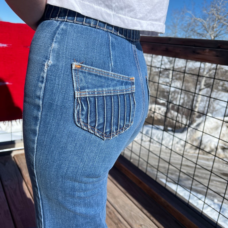 May include: A pair of blue denim jeans with a unique back pocket design featuring a series of vertical stitched lines. The jeans are fitted and have a high waist.