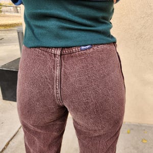 Pode incluir: Close-up de jeans de ganga cor de vinho. Os jeans t&ecirc;m um design cl&aacute;ssico de cinco bolsos, fecho de bot&atilde;o e z&iacute;per e uma etiqueta Wrangler no bolso de tr&aacute;s. O tecido tem um aspeto ligeiramente desbotado.