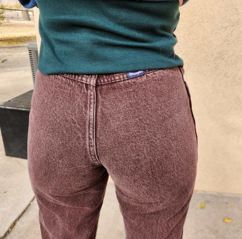 Pode incluir: Close-up de jeans de ganga cor de vinho. Os jeans t&ecirc;m um design cl&aacute;ssico de cinco bolsos, fecho de bot&atilde;o e z&iacute;per e uma etiqueta Wrangler no bolso de tr&aacute;s. O tecido tem um aspeto ligeiramente desbotado.