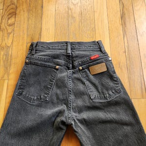 May include: Close-up of the back of black Wrangler jeans. The jeans have two back pockets, a red Wrangler tag, and a brown leather patch with the Wrangler logo. The jeans are made of a dark denim material.