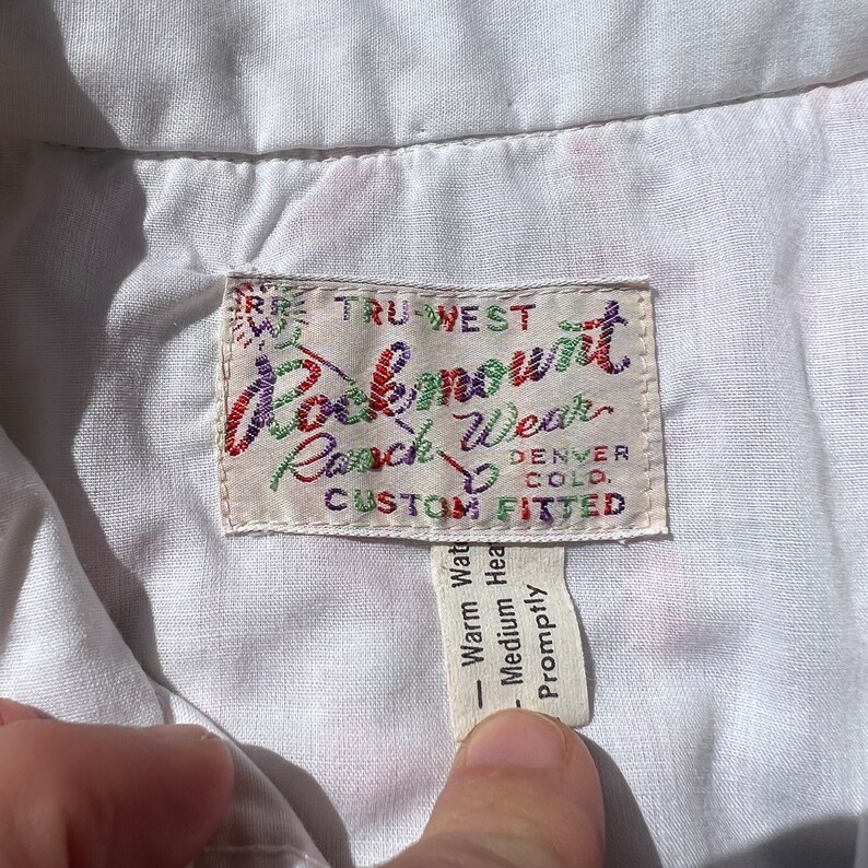 May include: A white label with the text "TRU-WEST Rockmount Ranch Wear DENVER COLO. CUSTOM FITTED" in colorful embroidery. The label is attached to a white shirt.