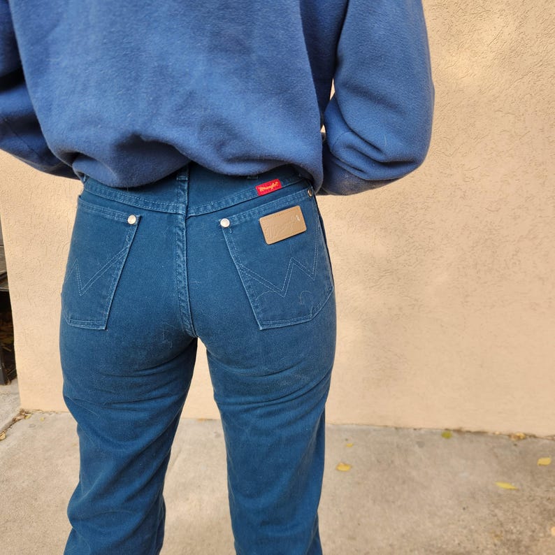 May include: Dark blue denim jeans with a high waist and a classic five-pocket design. The back pockets feature a signature stitching pattern. A brown leather patch and a red tag are visible on the back of the jeans.