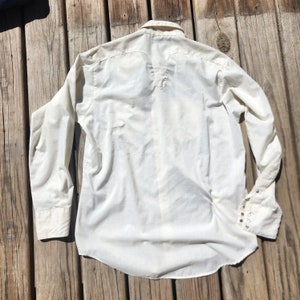 May include: Off-white long-sleeve shirt with a button-down front and cuffs. The shirt has a classic collar and a yoke detail on the back. The cuffs have multiple small silver-colored buttons. The shirt is made of a lightweight fabric.