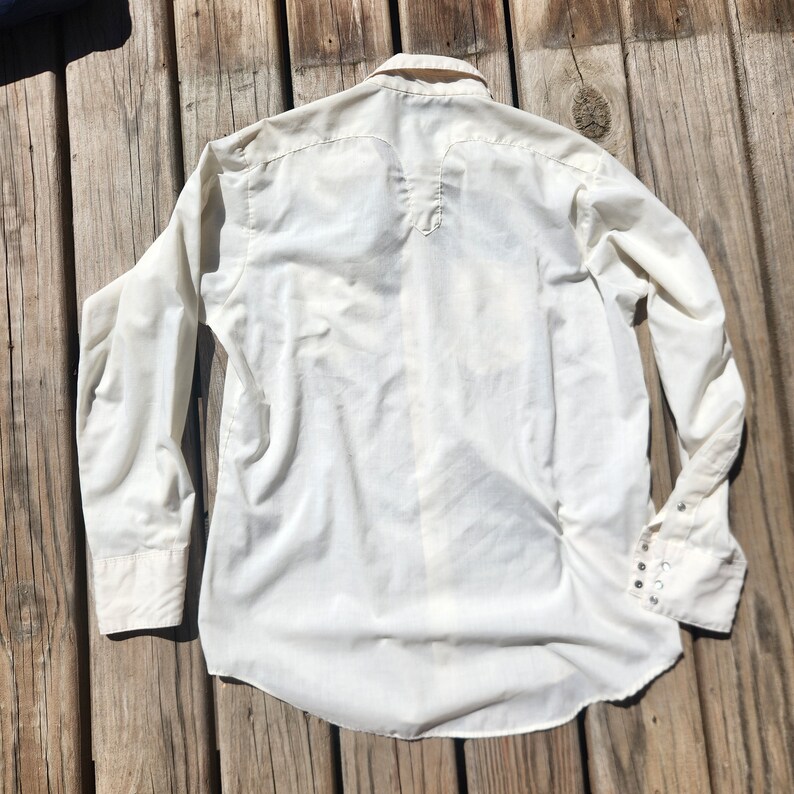 May include: Off-white long-sleeve shirt with a button-down front and cuffs. The shirt has a classic collar and a yoke detail on the back. The cuffs have multiple small silver-colored buttons. The shirt is made of a lightweight fabric.