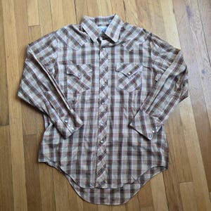 Puede incluir: Una camisa de manga larga con botones y un patrón de cuadros en tonos beige, marrón y blanco. La camisa incluye un cuello puntiagudo, dos bolsillos en el pecho con solapas puntiagudas y botones de presión plateados en el frente y los puños.