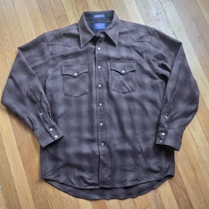 Peut inclure: Une chemise à manches longues à carreaux marron avec deux poches de poitrine et des fermetures à boutons-pression nacrés. La chemise a un col pointu et une fermeture boutonnée. L'étiquette Pendleton est visible sur le col.