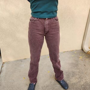 Pode incluir: Um par de jeans cor de vinho com um design cl&aacute;ssico de cinco bolsos. Os jeans t&ecirc;m um corte de perna reto e um fecho de bot&atilde;o e z&iacute;per. A pessoa tamb&eacute;m est&aacute; usando uma camisa verde escura e tamancos azuis escuros.