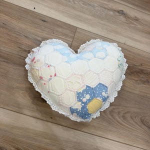 May include: A heart-shaped pillow constructed from a patchwork of hexagonal fabric pieces in various pastel colors and floral patterns. The pillow is edged with a white eyelet lace trim, creating a decorative border around the entire heart shape.