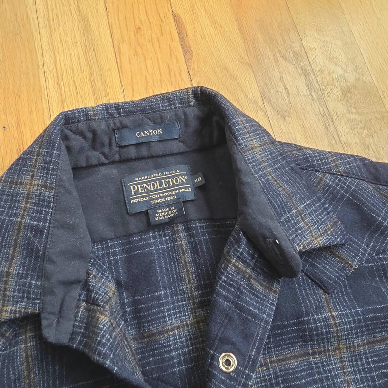 May include: A navy blue and brown plaid shirt with the word "CANYON" on a tag. The shirt has a button closure and a label that reads "PENDLETON WOOLEN MILLS SINCE 1963". The shirt is size XS and made in Mexico of USA fabric.