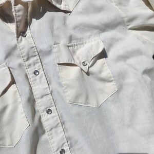 May include: Cream-colored Western-style shirt with two front pockets and snap closures. The shirt features a classic yoke design and pearl snap buttons. A vintage-inspired garment.
