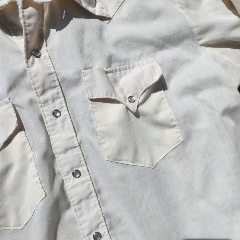 May include: Cream-colored Western-style shirt with two front pockets and snap closures. The shirt features a classic yoke design and pearl snap buttons. A vintage-inspired garment.