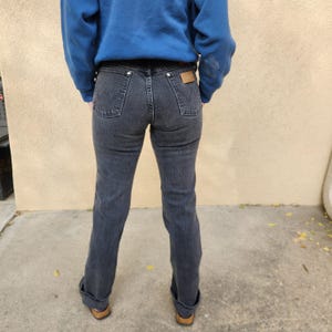 May include: Dark gray denim jeans with a classic five-pocket design. The jeans have a straight leg cut and are cuffed at the ankles. A brown leather patch is visible on the back waistband. The person is wearing a blue sweatshirt.
