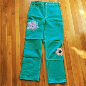 May include: Teal blue denim pants with two floral patches. The patches are made of pink, white, and green fabric.