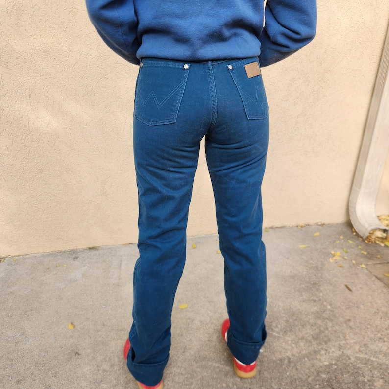 May include: Dark blue denim jeans with a high waist and a classic five-pocket design. The jeans have a straight leg cut and a small brown leather patch on the back. The person is wearing red shoes.