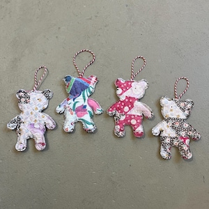 May include: Four handmade fabric bear ornaments with red and white string hangers. Each bear is made from patterned fabric in various colors, including pink, white, and floral designs. The bears are stitched together with white thread.