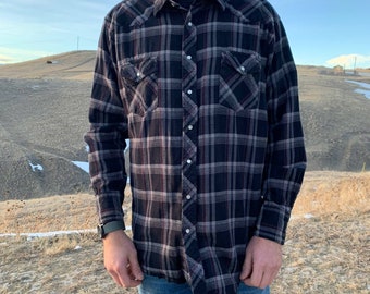 May include: A long-sleeved plaid shirt in black, gray, and burgundy. The shirt has a button-down front, two chest pockets, and a classic collar. The shirt is worn with blue jeans.