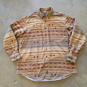 Puede incluir: Camisa abotonada de manga larga con un estampado de inspiración suroeste en tonos beige, marrón y morado. La camisa tiene cuello, bolsillo en el pecho y puños abotonados. Se muestra sobre una superficie neutra.