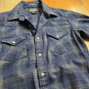 May include: A navy blue plaid button-up shirt with two chest pockets and silver snap closures. The shirt has a classic collar and the brand name "Pendleton" is visible on the label.