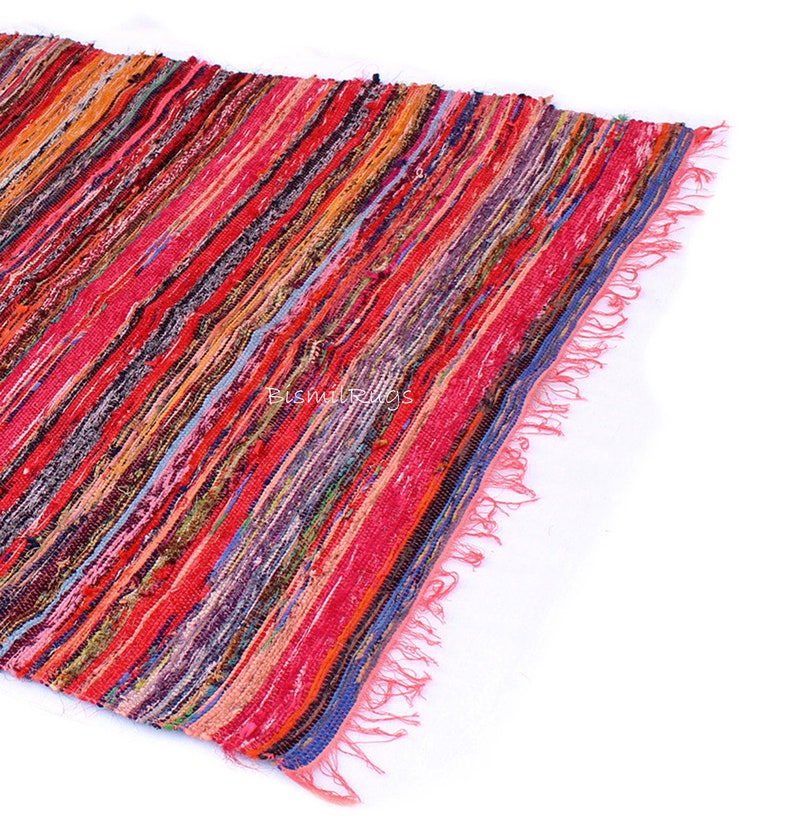 May include: A colorful, striped rug with a mix of red, orange, pink, and blue hues. The rug has a fringed edge and a woven texture. The rug is made of fabric strips and is suitable for home decor.