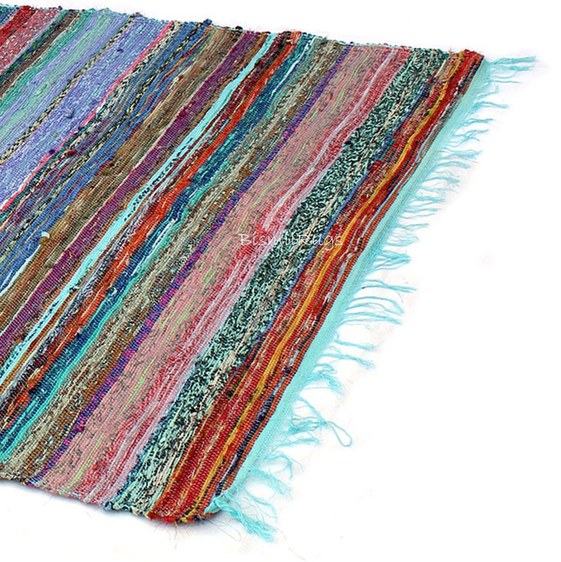 May include: A colorful, striped rag rug with a turquoise fringe. The rug features a variety of colors including red, blue, purple, and green. The rug is made of woven fabric.