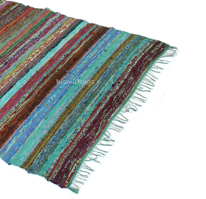 May include: A colorful, striped rag rug with a variety of colors including teal, burgundy, brown, and blue. The rug has a fringed edge and is made of woven fabric strips. The rug is angled, showing the diagonal stripes.