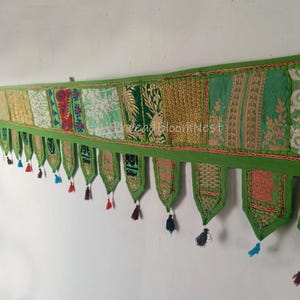 Puede incluir: Un tapiz de pared largo y horizontal hecho de tela con un mosaico de paneles bordados en verde, dorado y blanco. El borde inferior está rematado con varias pestañas de tela puntiagudas y colgantes, cada una adornada con una pequeña borla colorida.