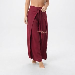 May include: Deep burgundy wrap pants featuring a high-waisted design with a long, integrated fabric tie at the center front. The garment has wide legs with an overlapping front panel that creates a split effect. The text TBN is visible.