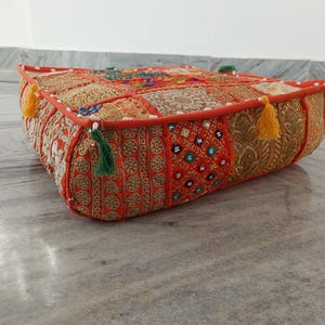 May include: A square floor cushion featuring a patchwork design of orange, gold, and red fabric panels. The surface is decorated with intricate embroidery, small reflective mirrors, and gold thread work. Yellow and green tassels hang from the corners.