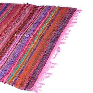 May include: A colorful, striped rug with a pink fringe. The rug features a variety of colors, including pink, purple, orange, and green. The rug is made of woven fabric and has a textured appearance. The text "BishnuRugs" is visible on the rug.