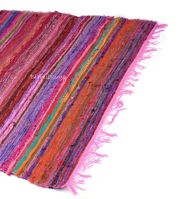 May include: A colorful, striped rug with a pink fringe. The rug features a variety of colors, including pink, purple, orange, and green. The rug is made of woven fabric and has a textured appearance. The text "BishnuRugs" is visible on the rug.