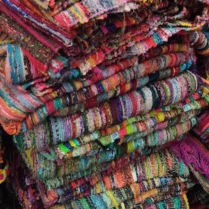 May include: A stack of colorful, woven rag rugs. The rugs display a variety of colors including pink, red, yellow, green, and blue. The rugs appear to be made of strips of fabric woven together, creating a textured surface.
