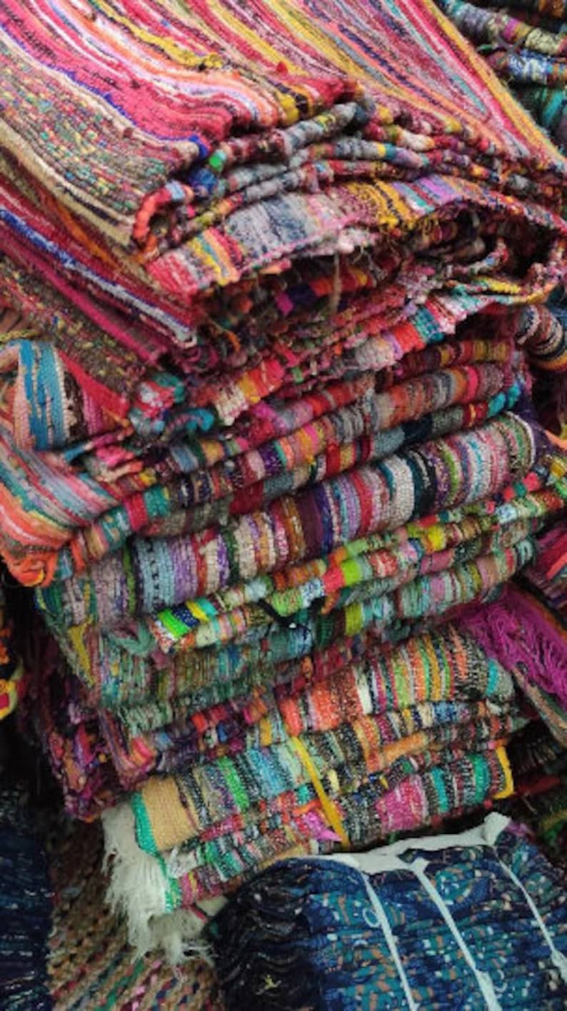 May include: A stack of colorful, woven rag rugs. The rugs display a variety of colors including pink, red, yellow, green, and blue. The rugs appear to be made of strips of fabric woven together, creating a textured surface.