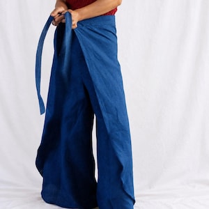 May include: A pair of deep blue wrap-style trousers featuring a high waistband and a long, integrated fabric tie that hangs down from the front. The pants have a wide-leg silhouette and are constructed from a solid-colored, textured fabric.